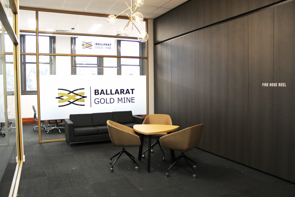 Modern office waiting area with 'Ballarat Gold Mine' branding on the wall, a dark sofa, two tan-colored chairs, and a coffee table.