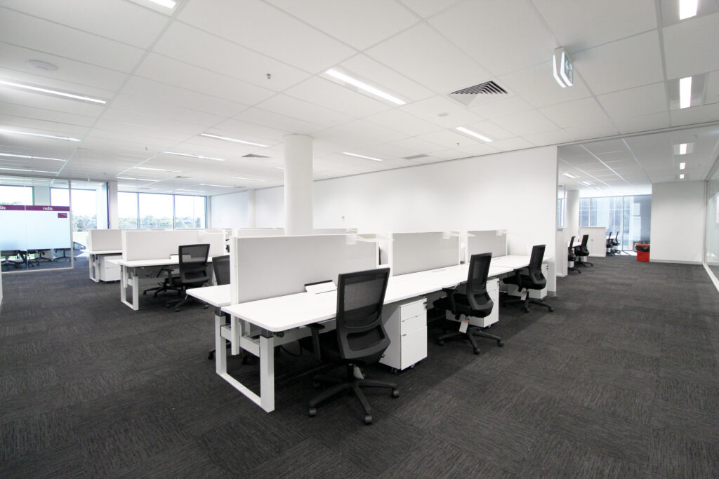 Spacious open-plan office space with white desks, ergonomic chairs, and 'ndis' branding, illuminated by overhead lighting against a backdrop of large windows.