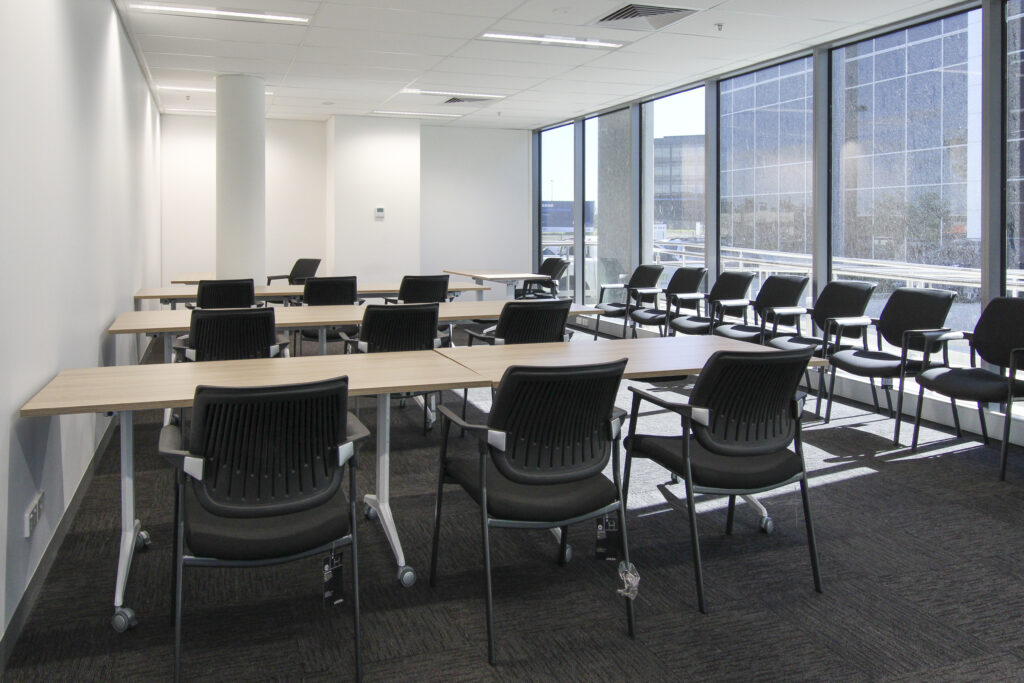 Spacious conference room with modern design, featuring light wooden tables, ergonomic black chairs, and large floor-to-ceiling windows offering an urban view.