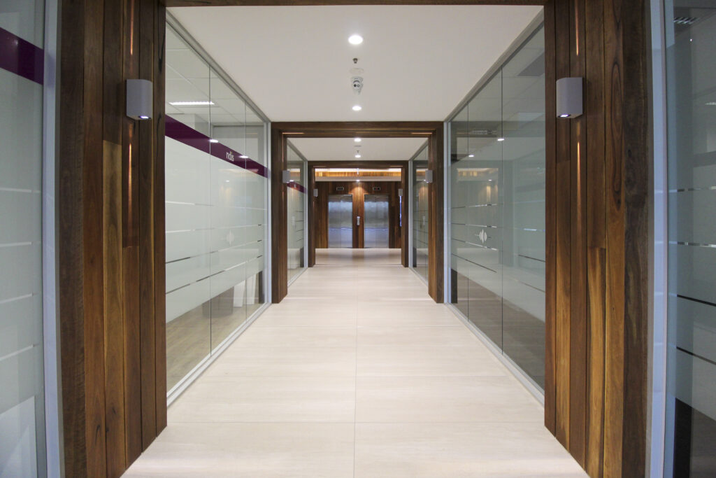 Elegant office corridor featuring wooden doors, glass partitions with 'ndis' branding, and modern lighting fixtures.