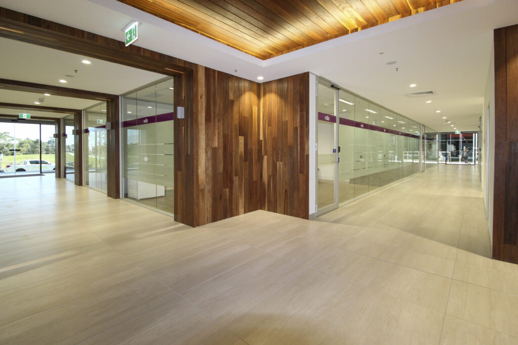 Modern office corridor with rich wooden accents, glass partitions displaying 'ndis' branding, and a light tiled floor leading to a panoramic window view.