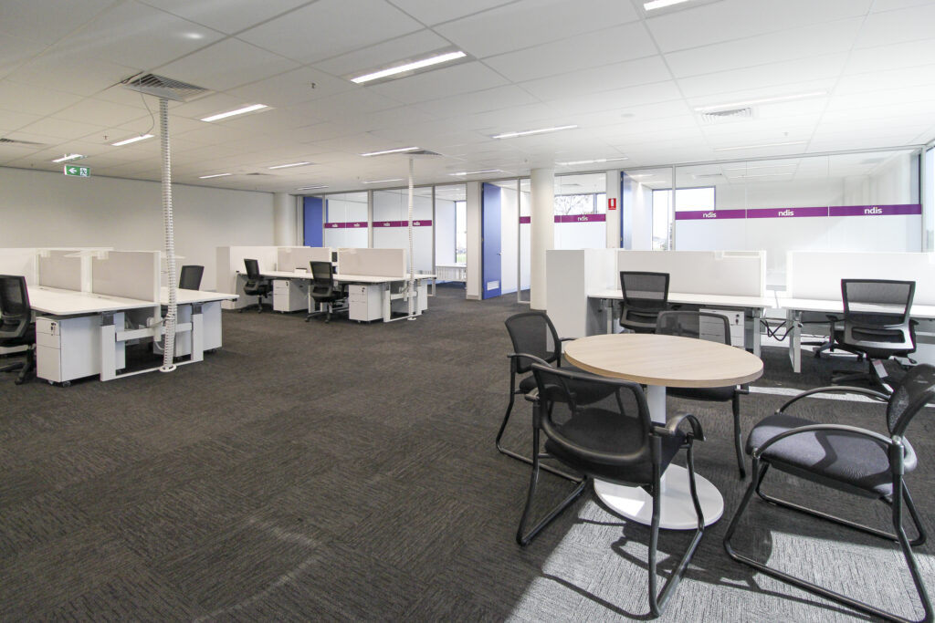 Spacious open-plan office space with individual workstations, ergonomic chairs, and meeting area, highlighted by 'ndis' branded partitions.