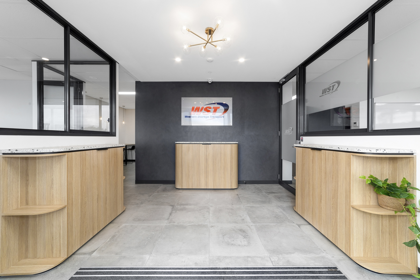 Contemporary office reception area featuring wooden countertops, a charcoal accent wall with a "WST - Western Storage Transport" logo, a chic gold chandelier, and clear glass dividers.